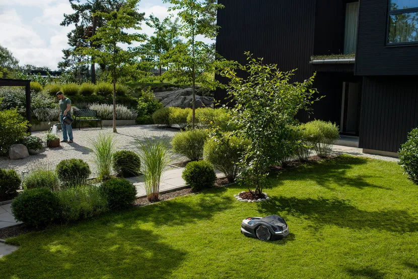 手入れの行き届いた庭で、背景に植物の世話をする人がいるロボット芝刈り機。